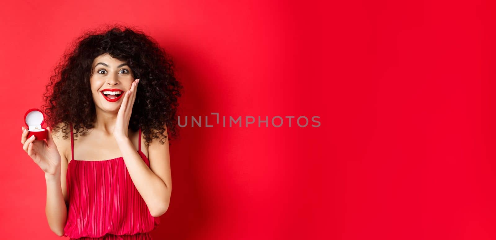 Image of surprised young woman with curly hairstyle, wearing red dress and lipstick, showing engagement ring, going to get married, standing on studio background.