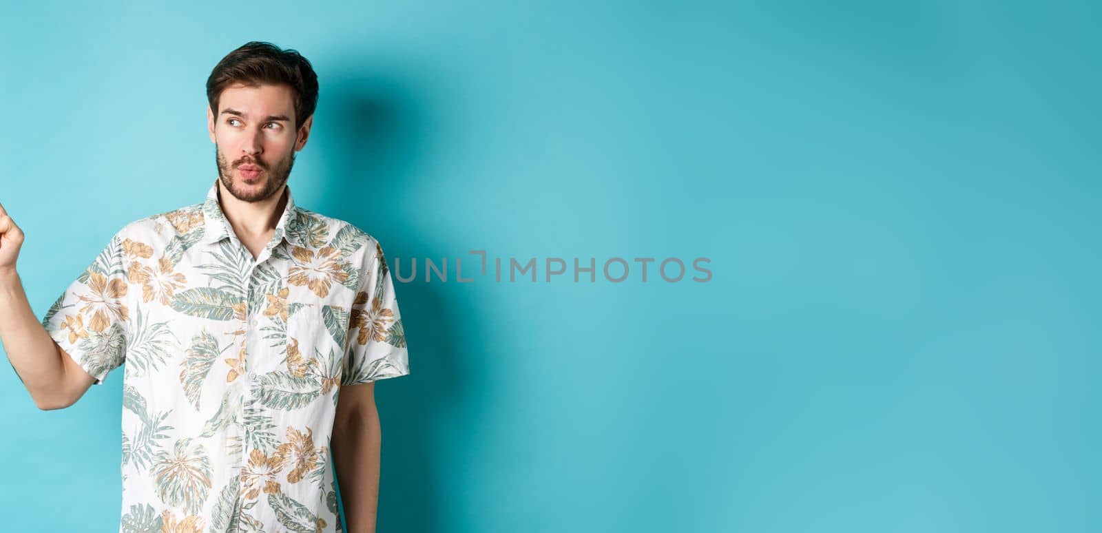 Handsome guy wearing summer shirt on vacation, pointing and looking aside at empty space, making amazed face, standing on blue background.