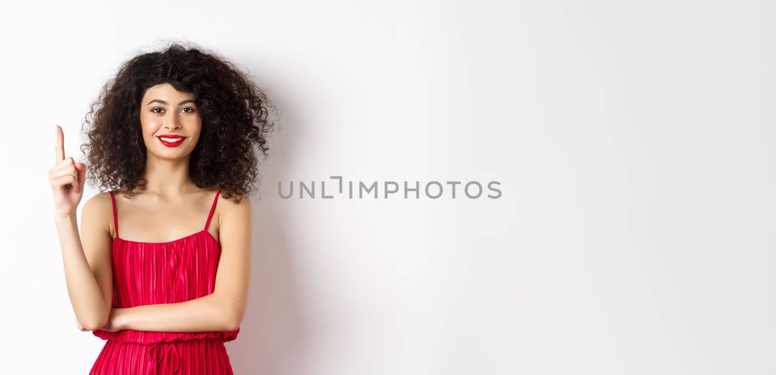 Beautiful smiling lady in red dress showing number one, raising finger and looking pleased, standing over white background.
