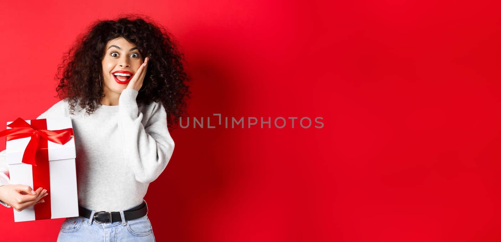 Surprised pretty girl receive Valentines day romantic gift, holding present box and looking with disbelief and happiness at camera, standing on red background by Benzoix