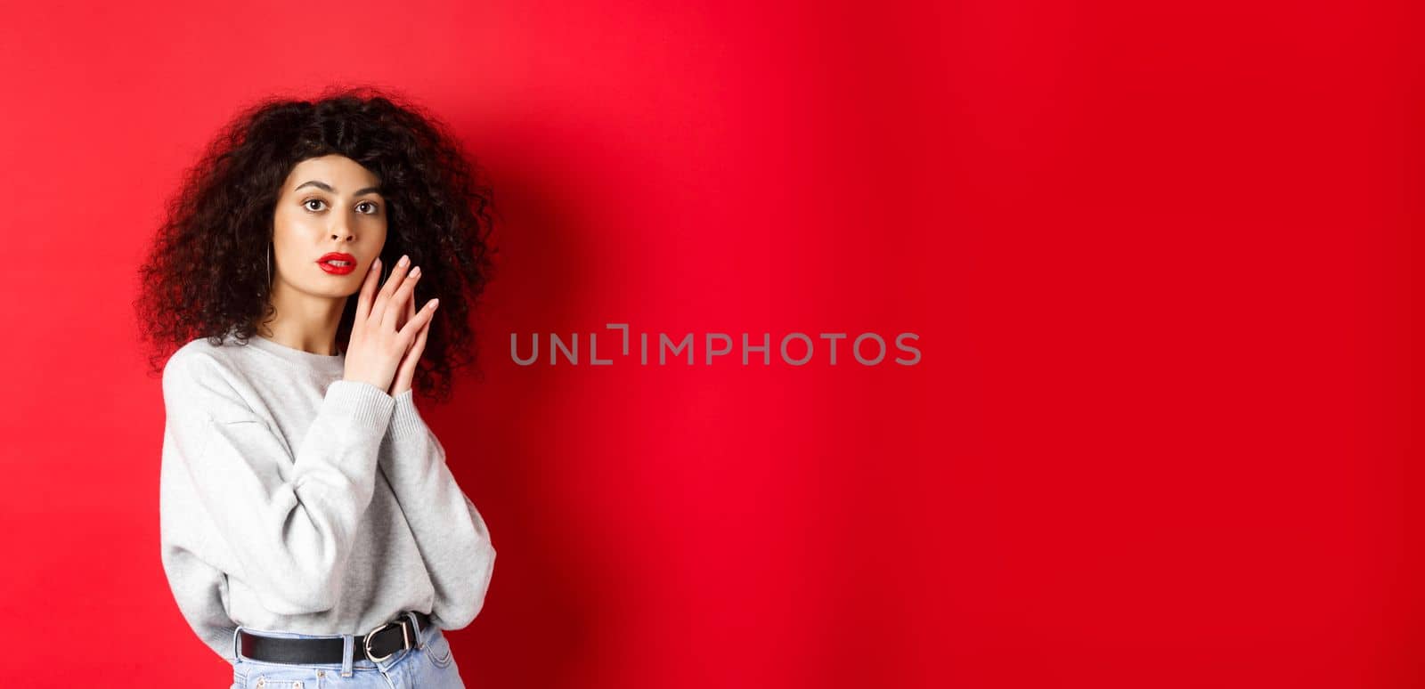 Sensual and romantic girl with curly hair and red lips, gently touching face with make-up, standing against red background.