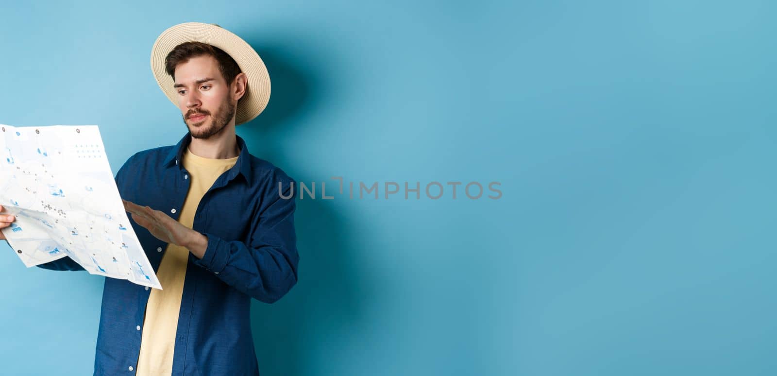 Image of handsome caucasian man looking at travel map, studying route during summer vacation, standing in straw hat on blue background.