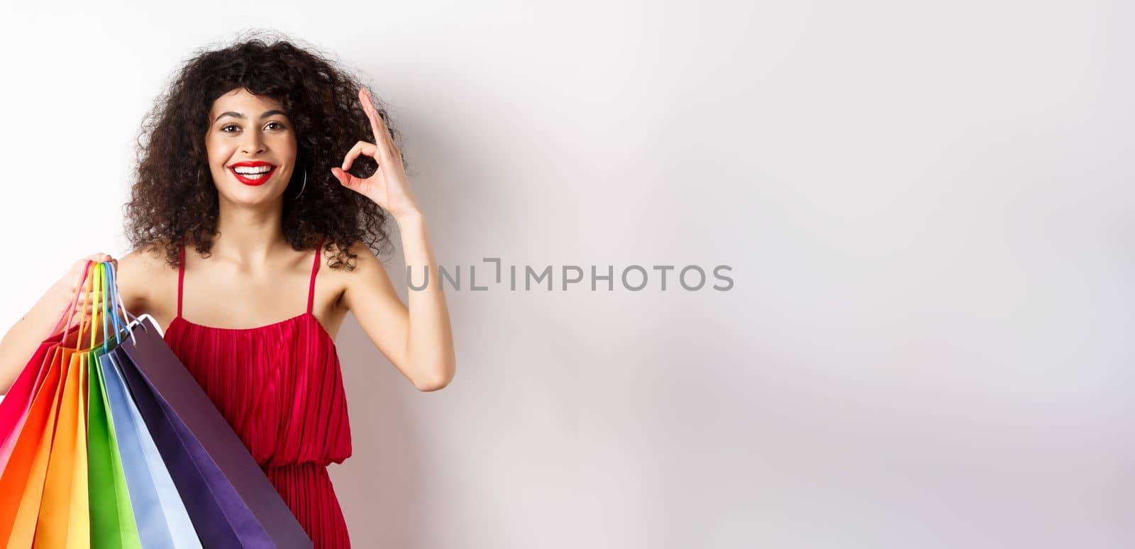 Beautiful woman in elegant dress, holding shopping bags and showing okay sign, recommend store with discounts, white background by Benzoix