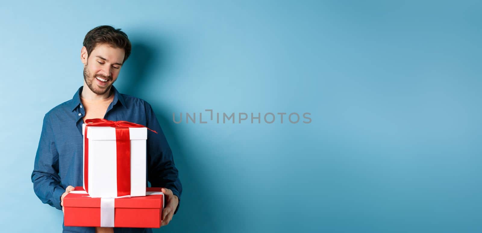 Romantic young man with beard, looking happy at gift boxes on valentines day, giving presents to lover, standing over blue background.