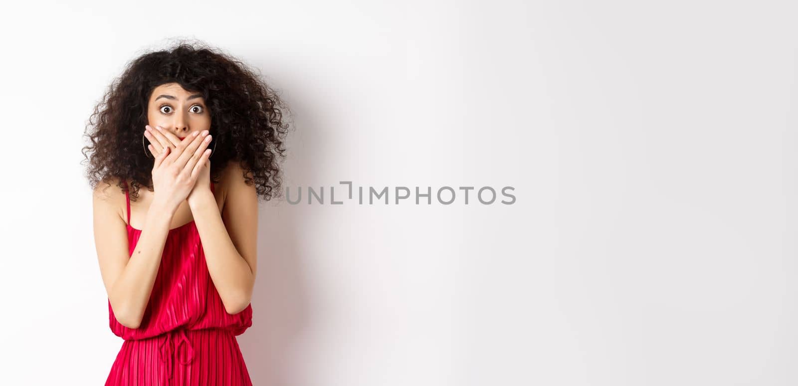 Shocked woman covering mouth with hands and staring with disbelief at camera, witness something shocking, standing in red dress on white background by Benzoix