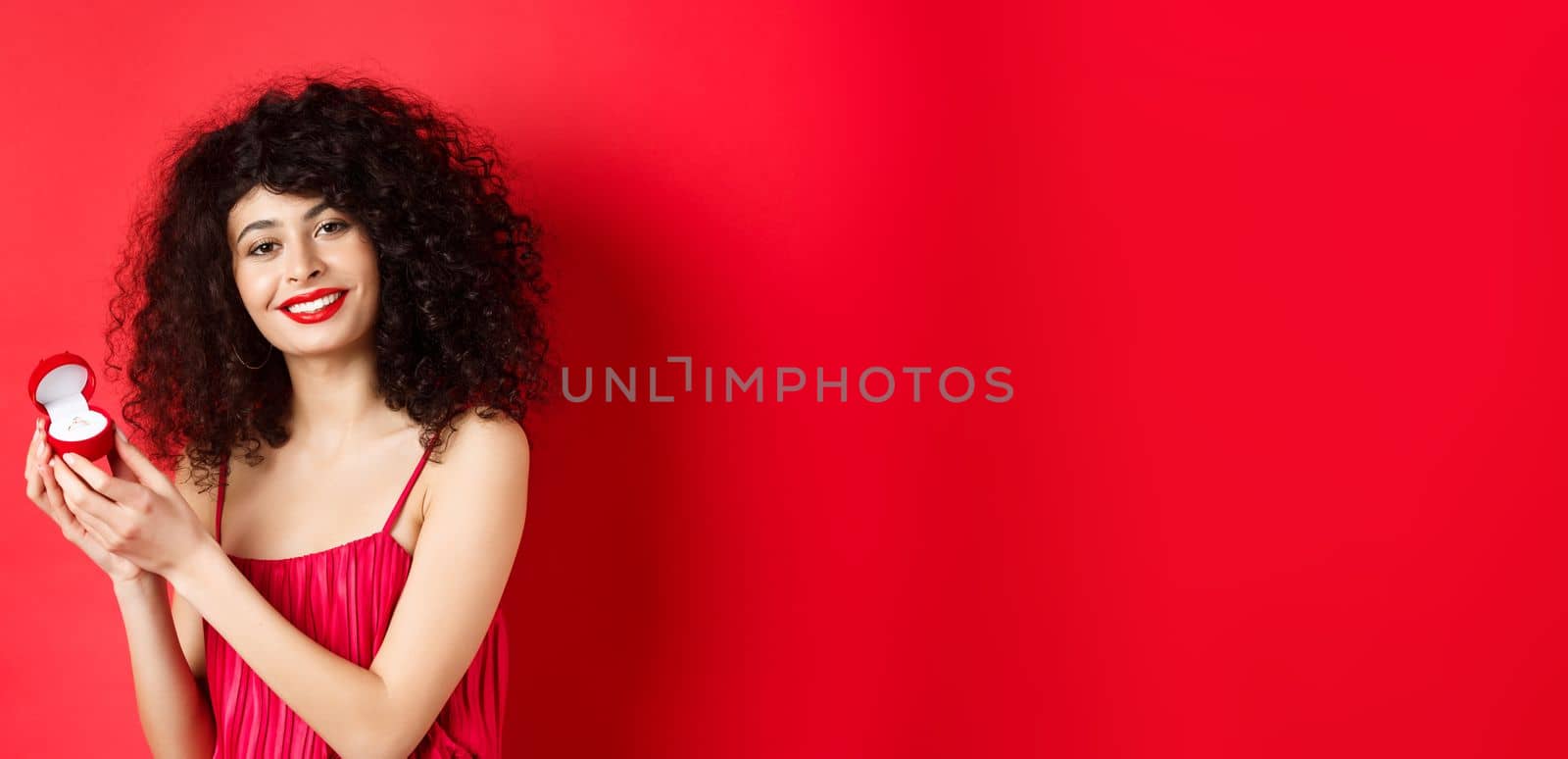 Romantic woman with curly hair, wearing red dress and showing engagement ring, standing happy on studio background.