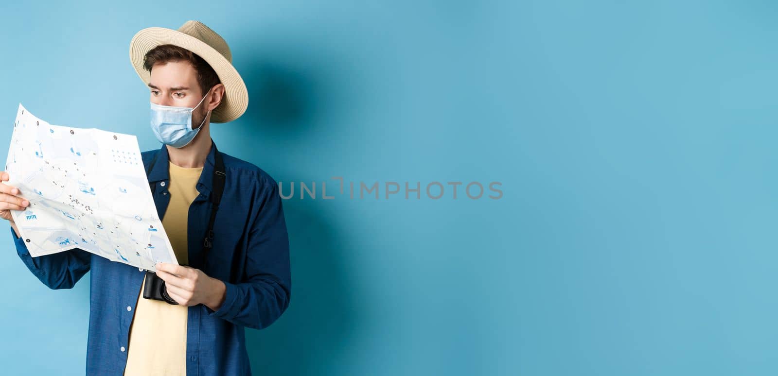 Covid-19, pandemic and travel concept. Handsome young guy in face mask and summer hat looking at tourist map, travelling on vacation, standing on blue background.