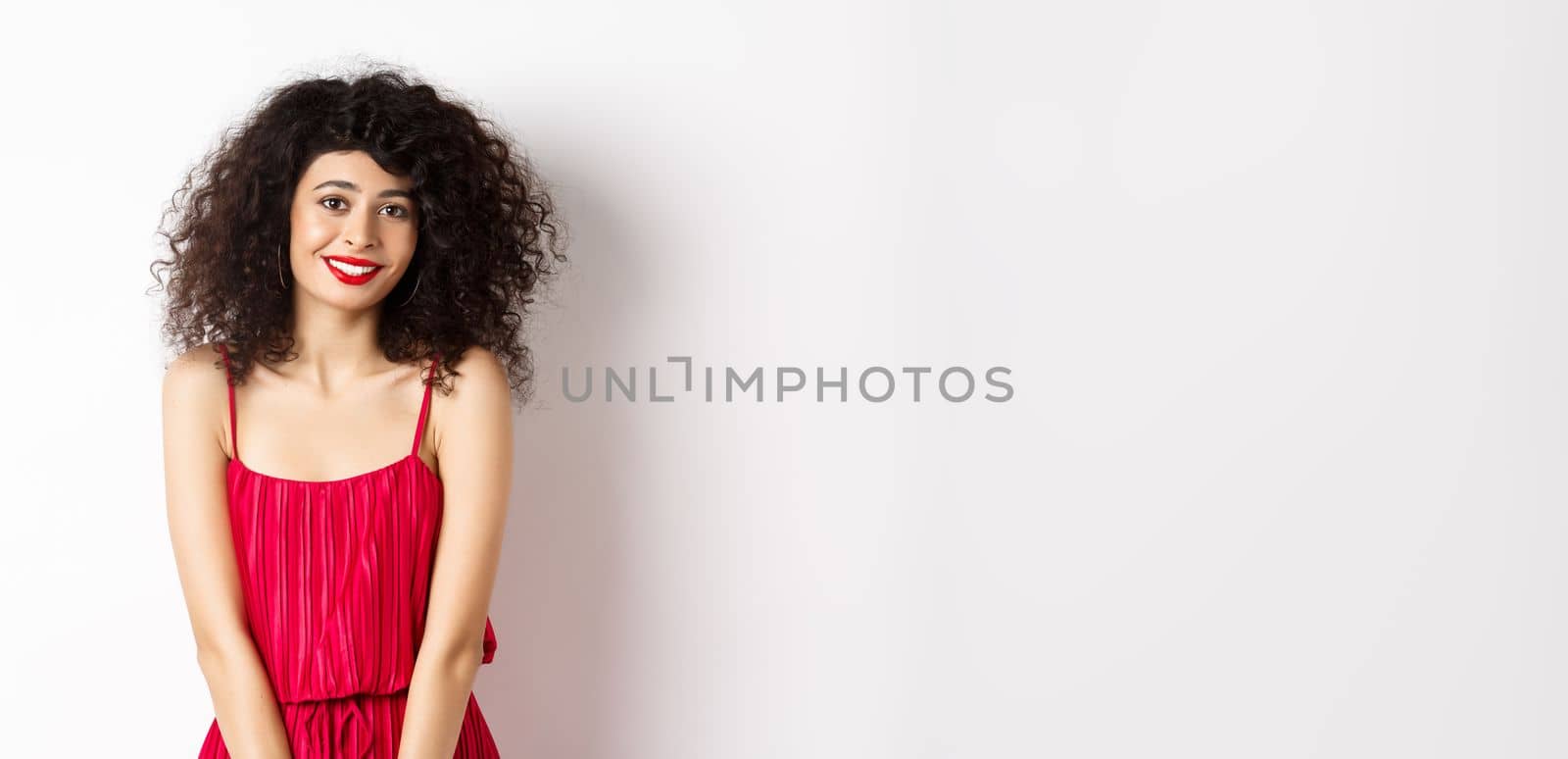 Cute modest lady in red dress and makeup, smiling and blushing, looking at camera, standing over white background.