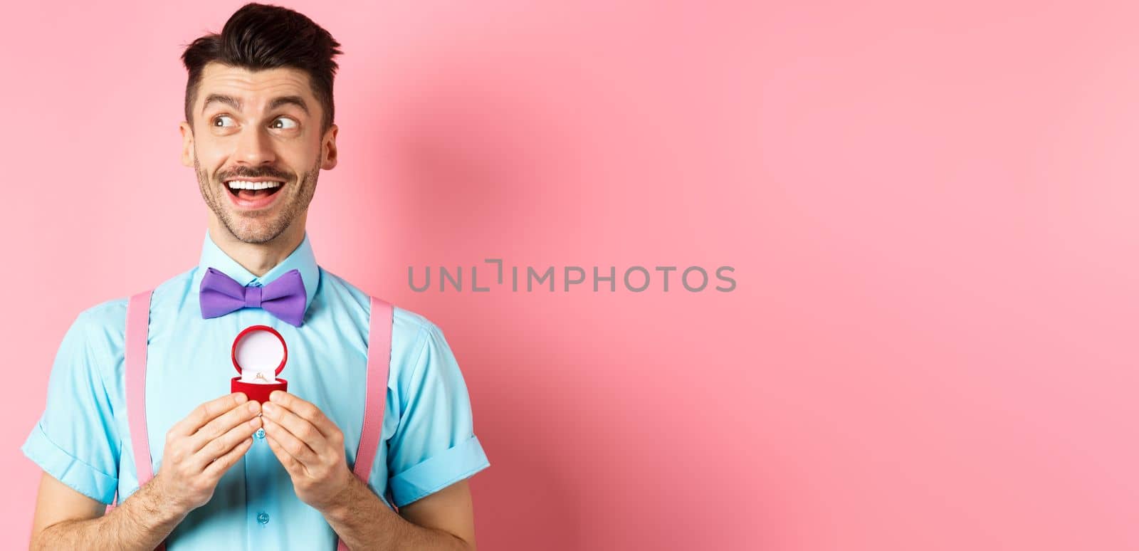 Valentines day. Romantic handsome man looking dreamy and smiling, showing engagement ring for his lover, standing happy over pink background by Benzoix