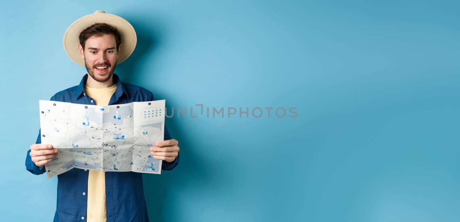 Cheerful tourist in summer hat planning travel route on vacation, looking at sighseeing map and smiling excited, standing on blue background.