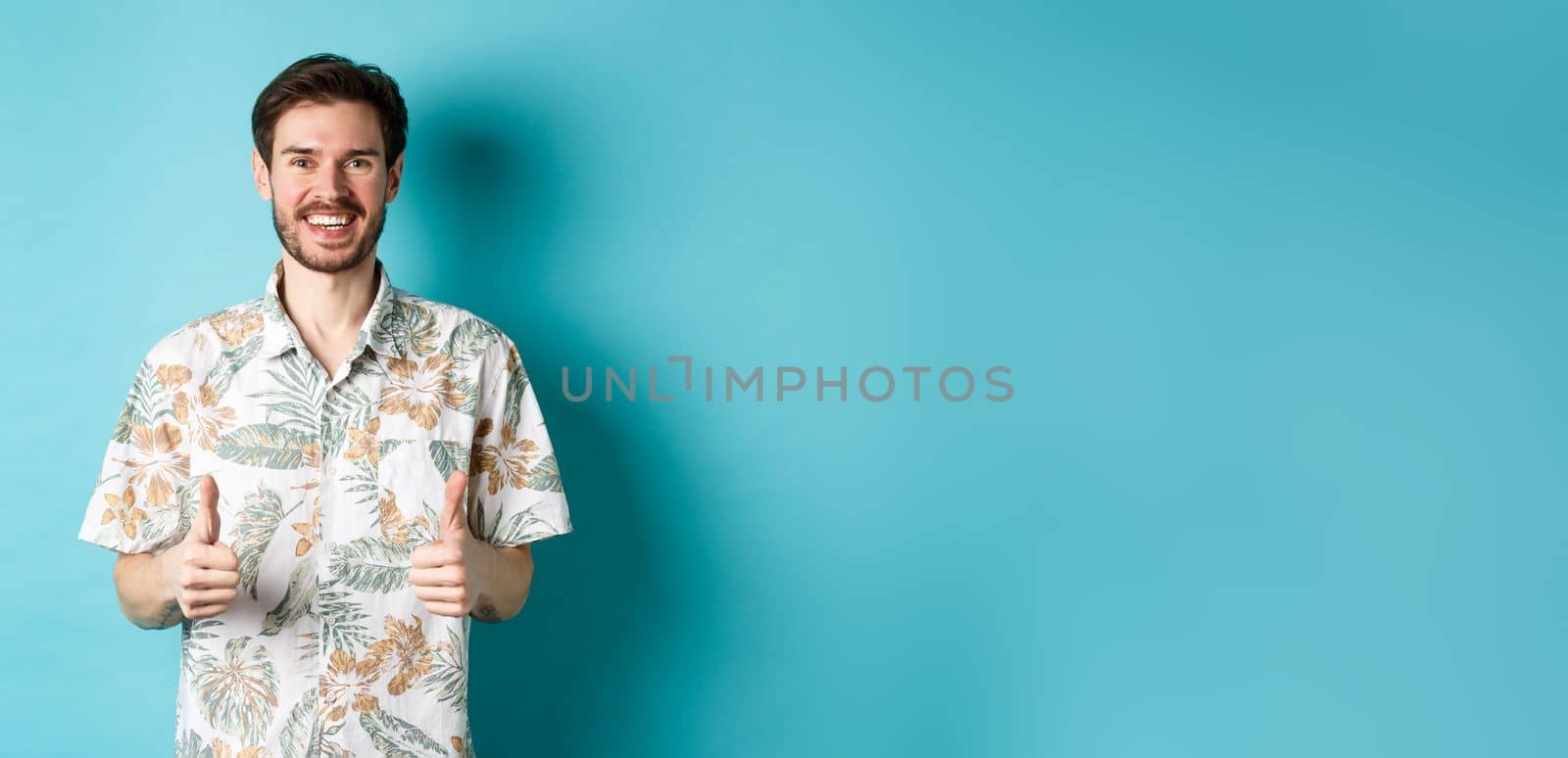 Summer holiday. Happy tourist showing thumbs up in approval and smiling, wearing hawaiian shirt, recommending travel agency, blue background.