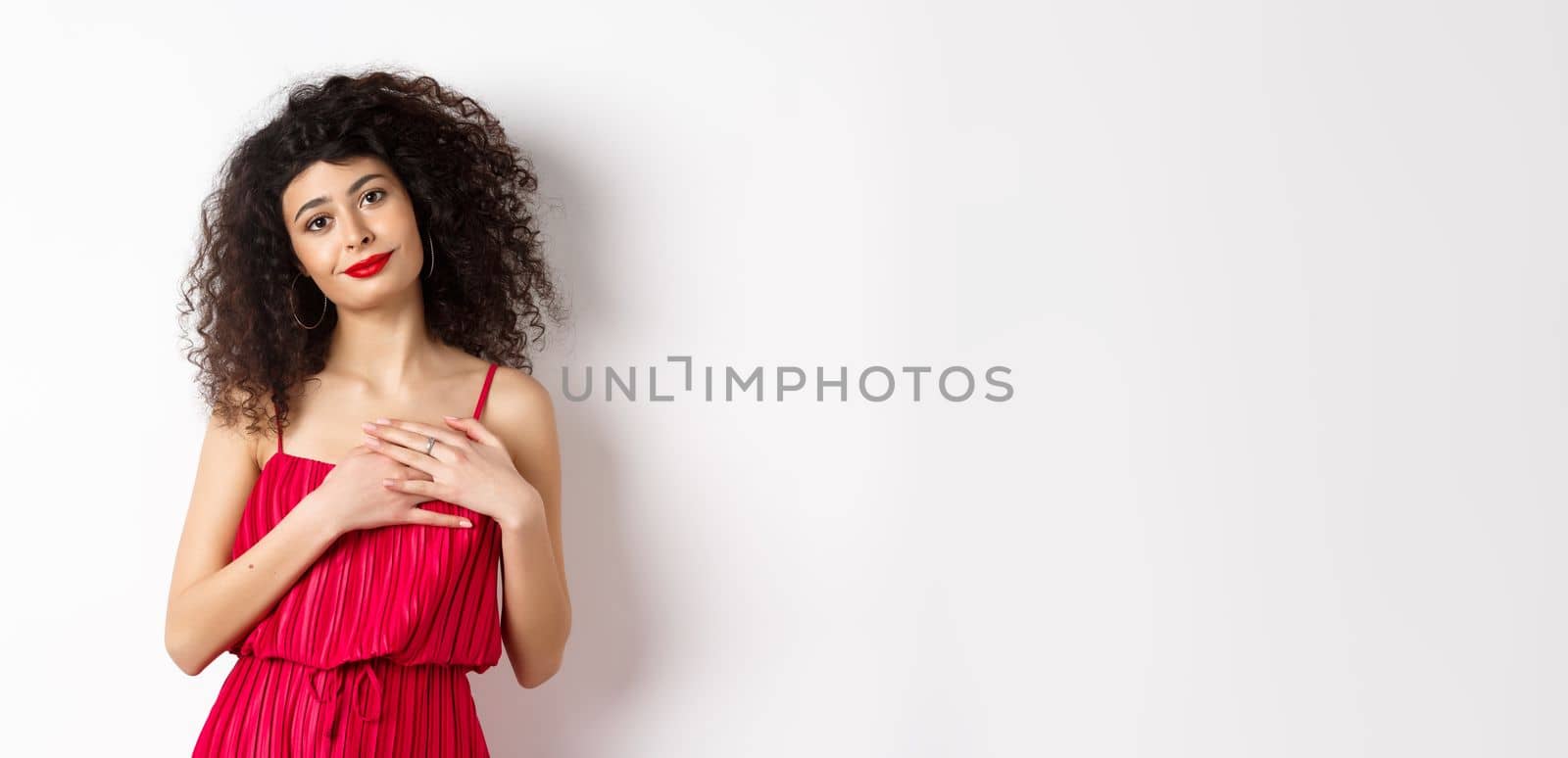 Touched young woman with curly hair, wearing red dress, holding hands on heart and smiling grateful, say thank you, standing over white background.