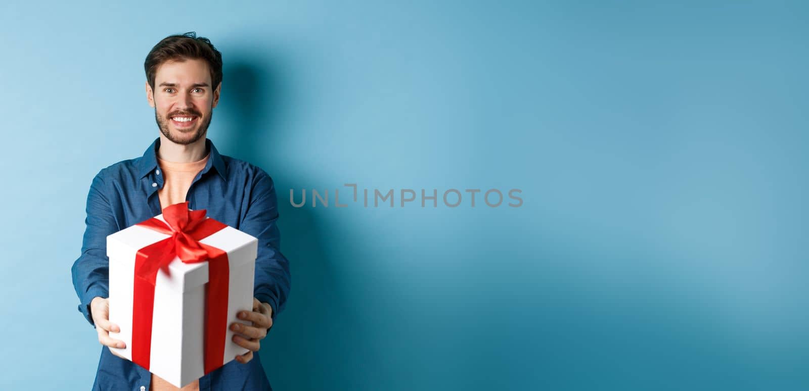 Valentines day. Handsome smiling man extending hands with gift box, wishing happy holiday. Guy making surprise present and looking cheerful, standing over blue background by Benzoix