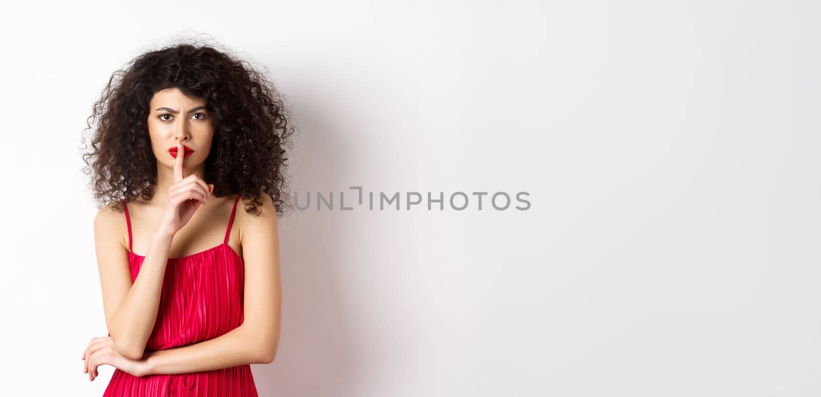 Angry elegant woman in red dress hushing and frowning, tell to be quiet, asking for silence, standing over white background.