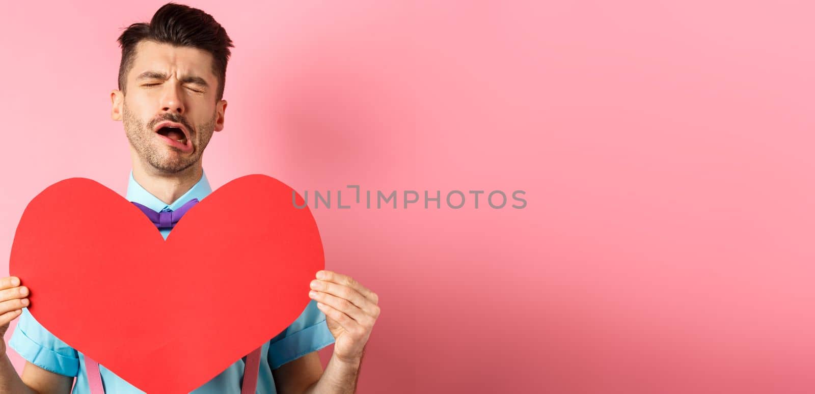 Valentines day concept. Sad and lonely man feeling heartbroken, being rejected, showing big red heart cutout and crying from break-up, standing on pink background.