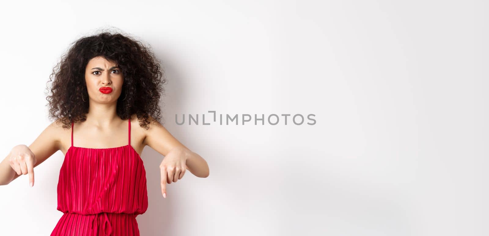 Disappointed caucasian woman in red dress complaining on spoiled date, frowning and grimacing, standing over white background.