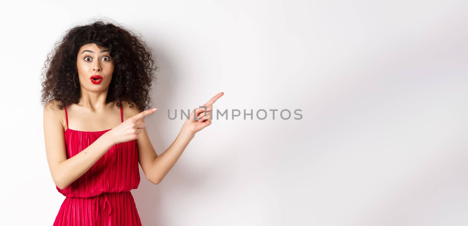 Surprised woman in red dress and makeup pointing fingers right, showing logo and look intrigued, standing on white background by Benzoix