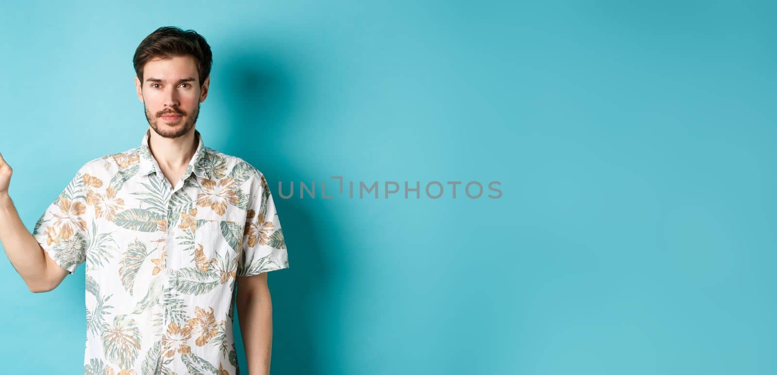 Portrait of handsome bearded guy on vacation, wearing summer shirt and pointing finger left at logo, standing on blue background.