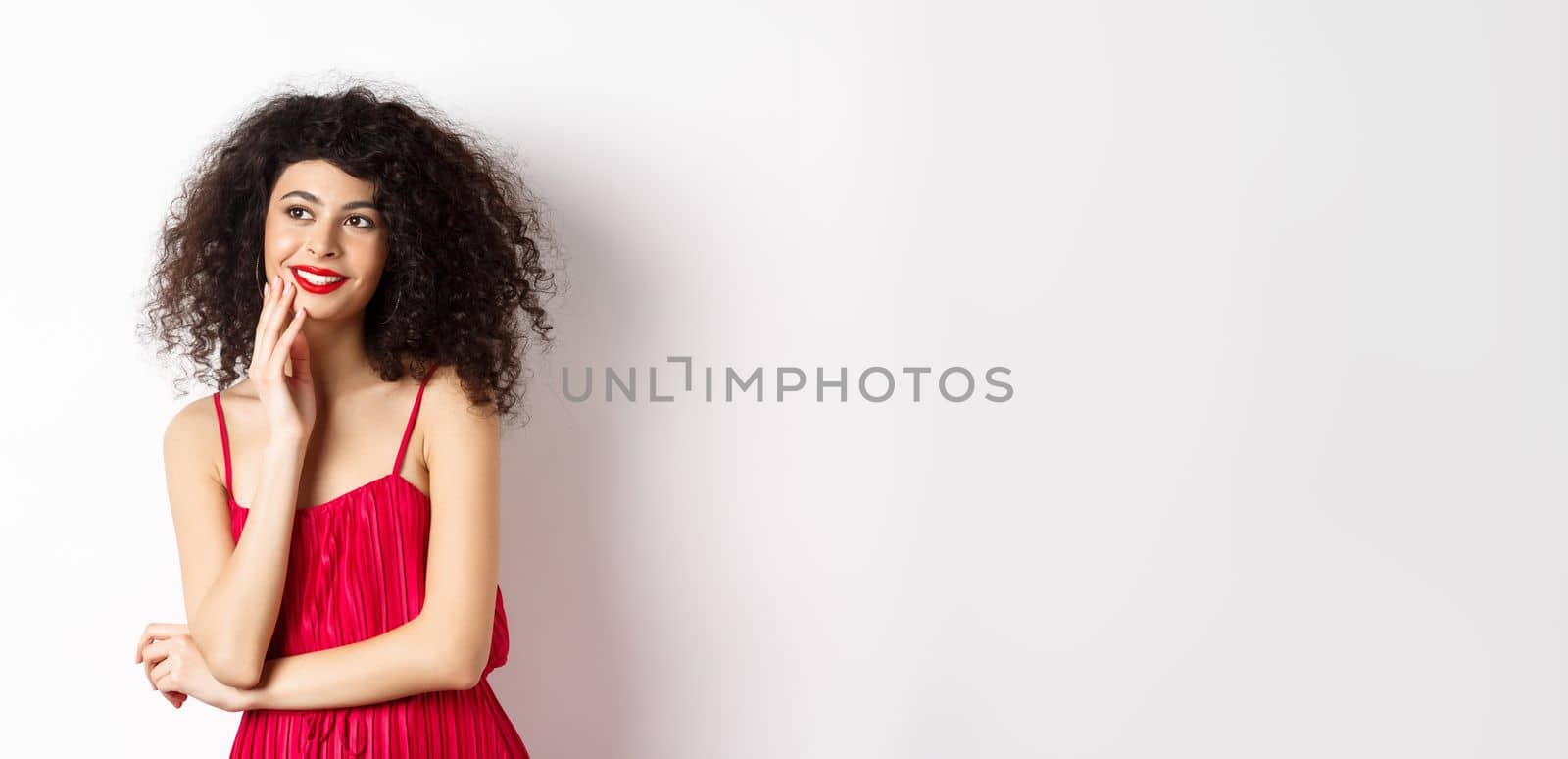 Dreamy elegant girl with curly hair, wearing red dress and makeup, looking left and smiling at logo, standing on white background.