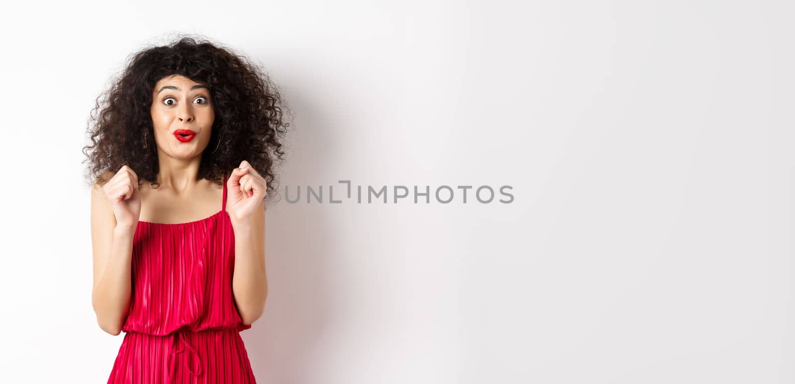 Excited smiling woman eager to try something, jumping from amusement and smiling, wearing red dress, standing on white background by Benzoix