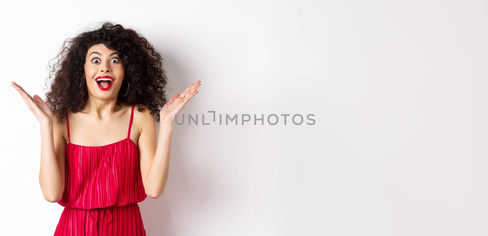 Surprised happy woman reacting to big news, looking cheerful with hands spread sidewas, standing in red dress on white background by Benzoix