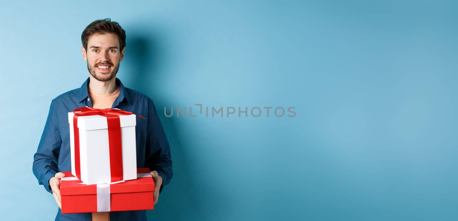 Happy valentines day. Handsome boyfriend holding gifts for lover on romantic date anniversary, standing over blue background.