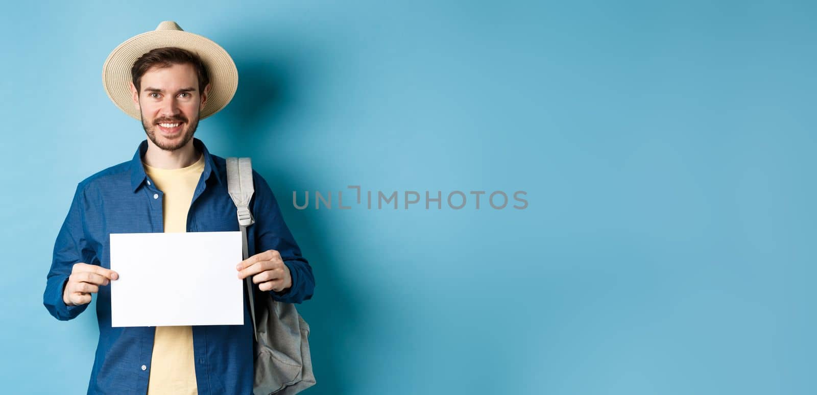 Happy tourist with backpack showing empty piece of paper, smiling at camera, standing on blue background. Concept of summer holiday and vacation by Benzoix