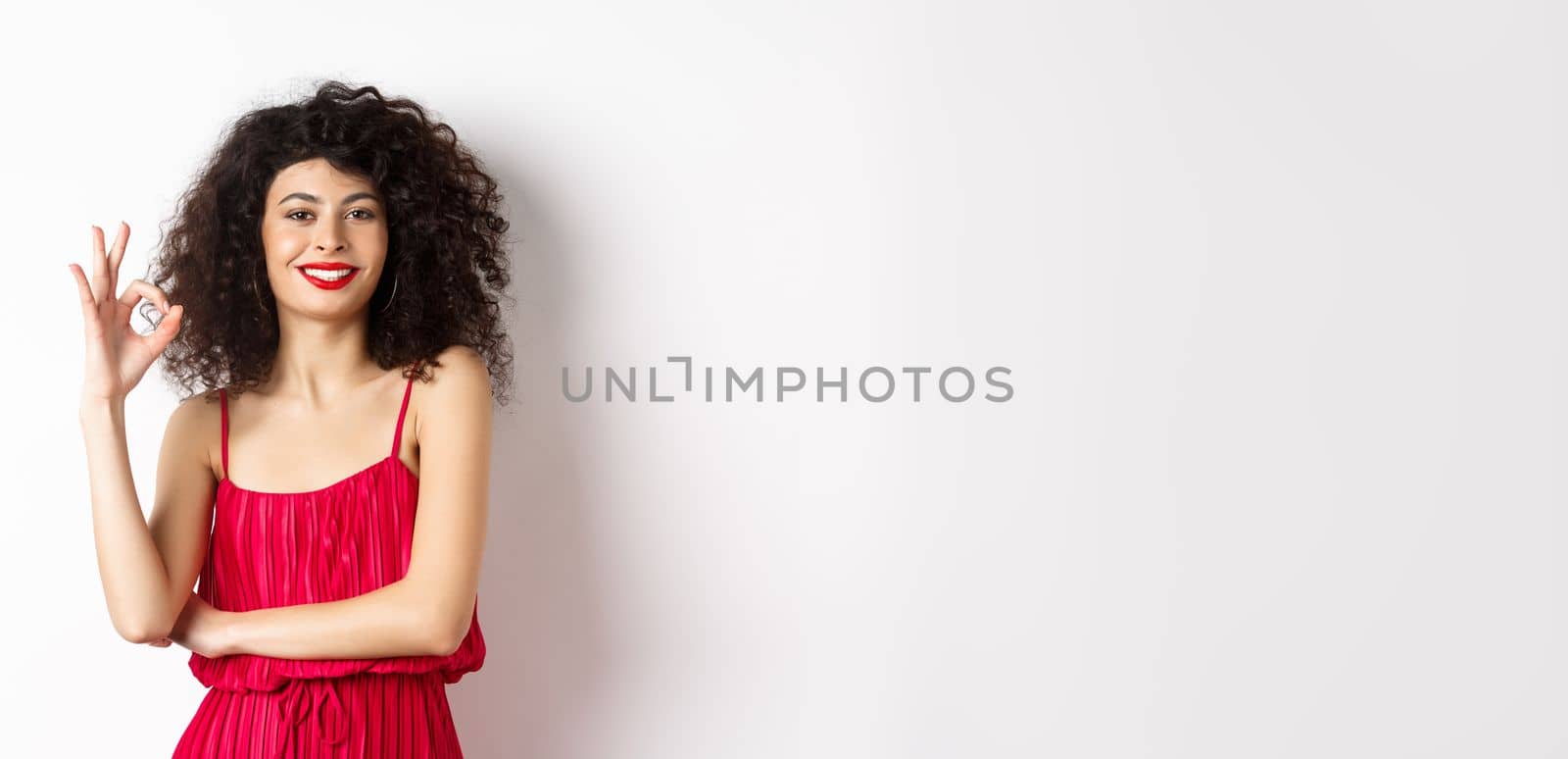 Elegant woman in romantic red dress and makeup, showing okay sign and smiling, recommending good thing, approve and agree, standing satisfied on white background.