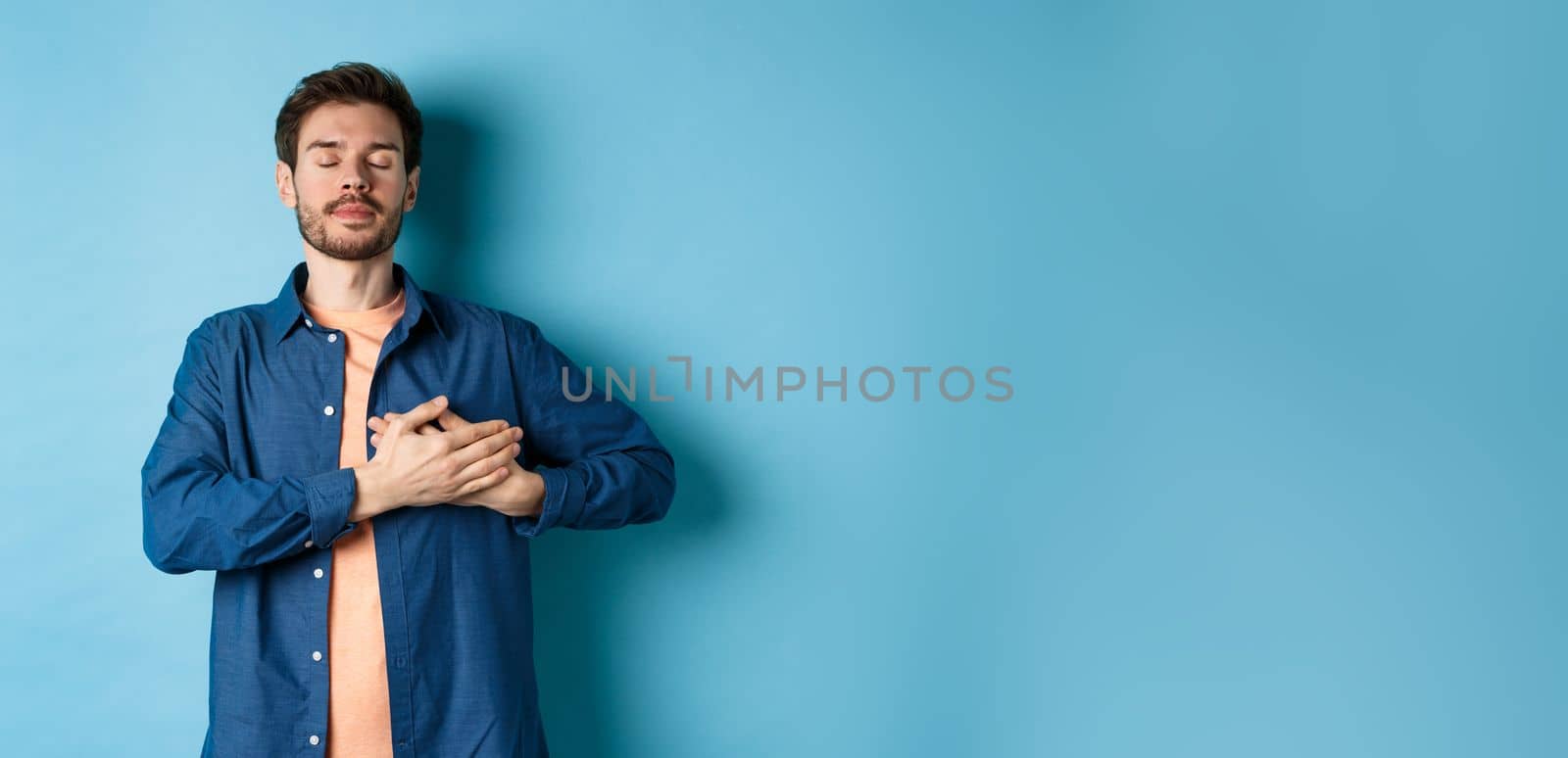 Romantic and nostalgic guy holding hands on heart with eyes closed, keep warm memories, standing on blue background.