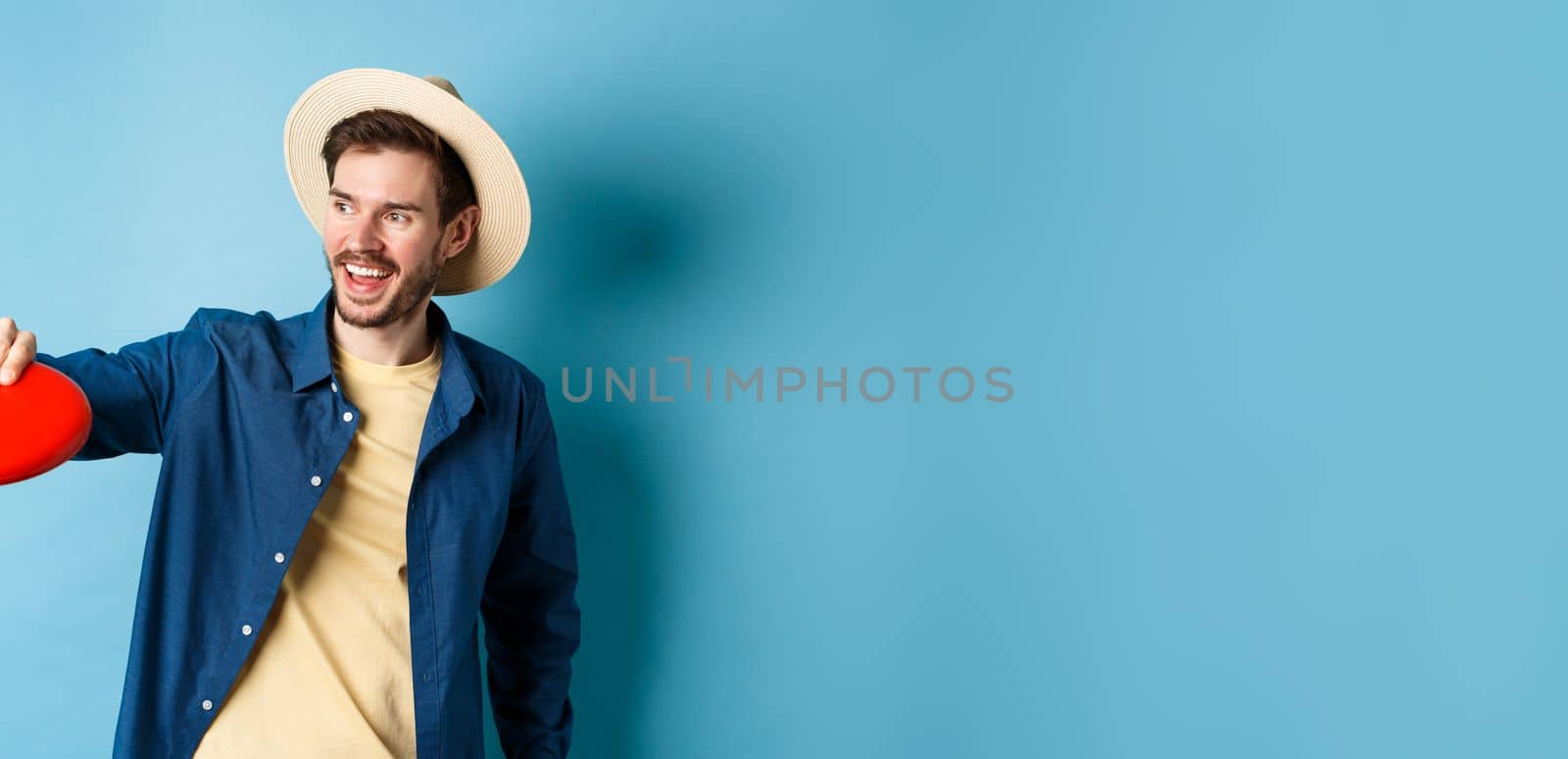 Happy smiling guy catching frisbee while playing with friends on summer vacation, standing in straw hat on blue background.