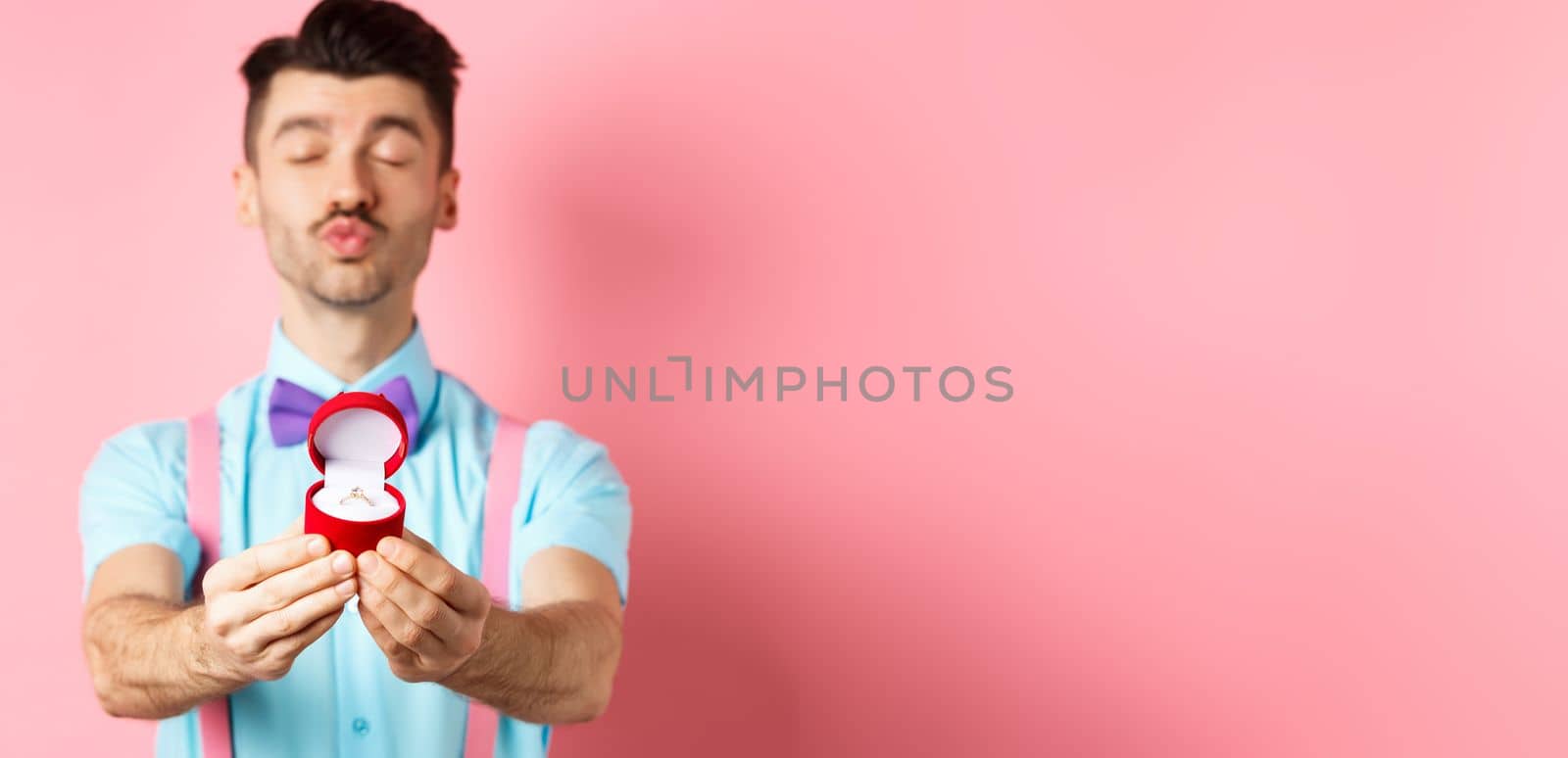 Valentines day. Silly boyfriend in bow-tie pucker lips, close eyes and waiting for kiss after making proposal, showing golden engagement ring in small red box, standing on pink background.