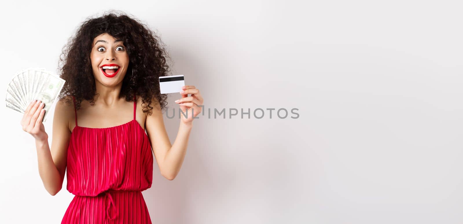 Happy attractive woman in red dress, screaming of joy and showing plastic credit card with money, winning prize, standing over white background by Benzoix