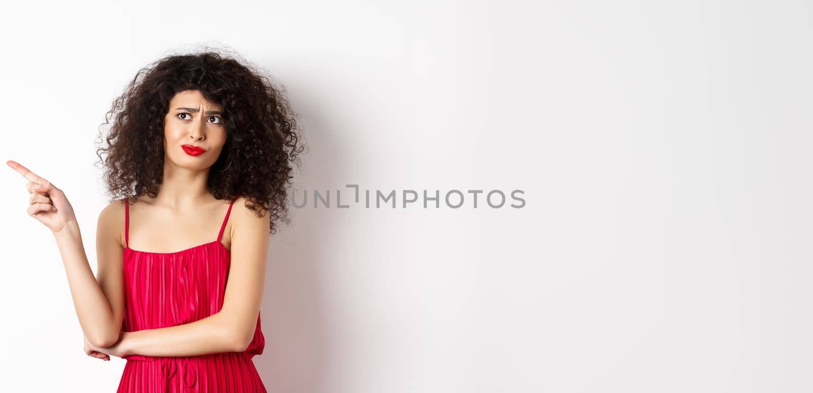 Disappointed and skeptical young woman with curly hair, wearing red dress, grimacing and pointing finger left at logo, standing over white background by Benzoix