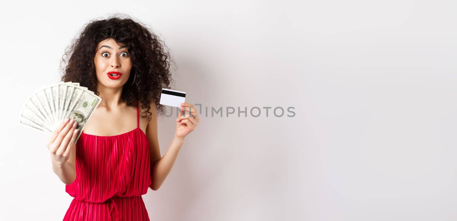 Excited woman looking with amazement and disbelief, showing dollars prize and plastic credit card, standing in red dress on white background.