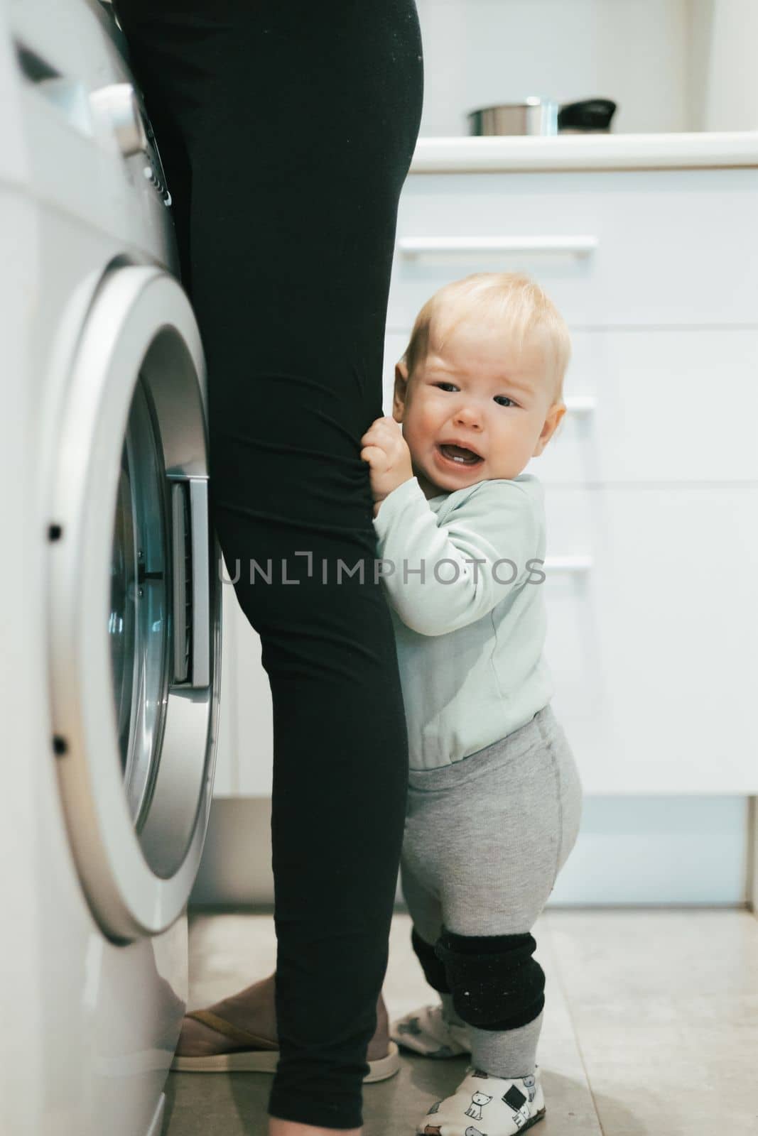 Little infant baby boy child hiding between mothers legs demanding her attention while she is multitasking, trying to do some household chores in kitchen at home. Mother on maternity leave