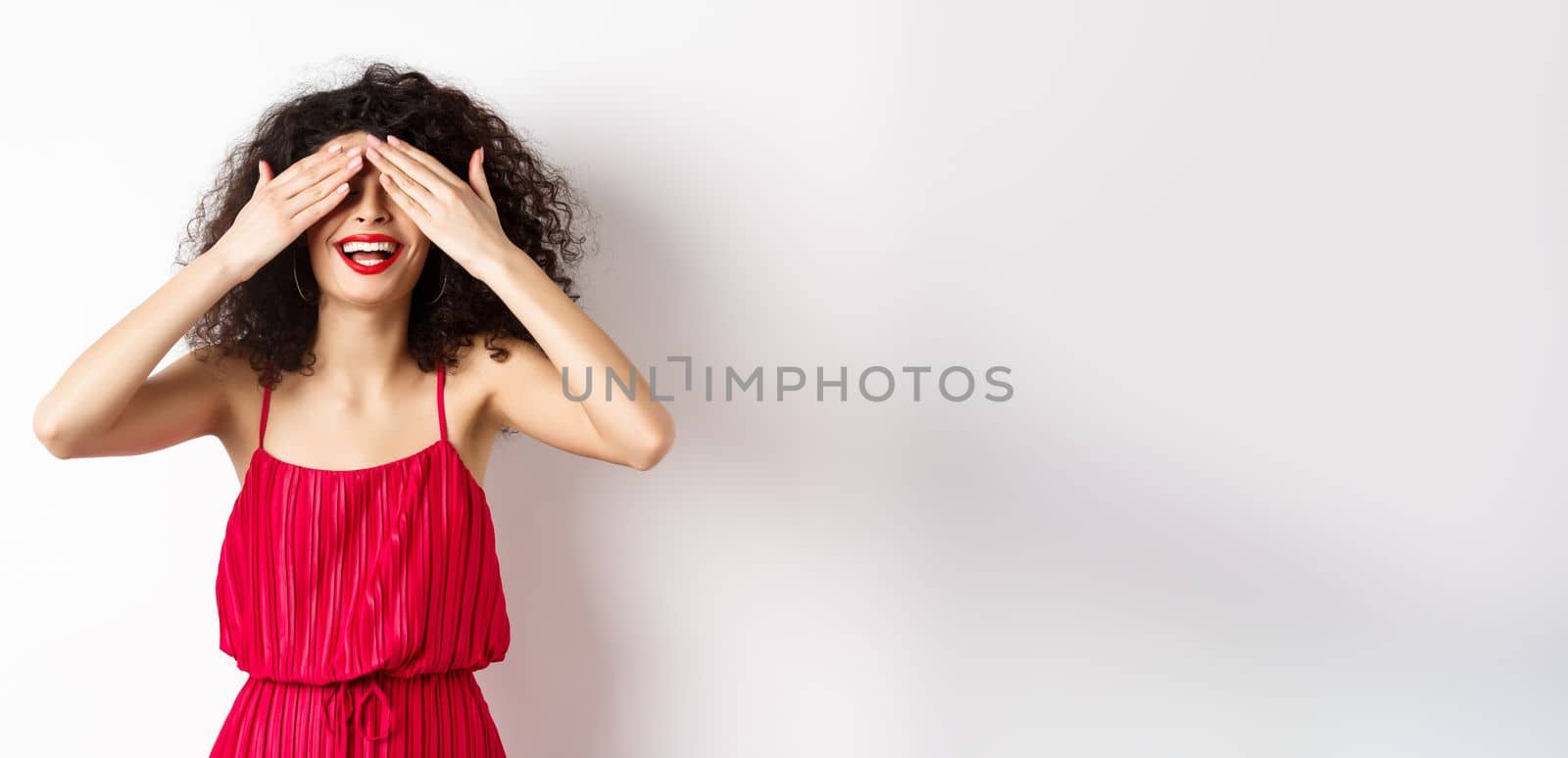 Excited curly-haired woman in party dress, close eyes and smiling with red lips, waiting for surprise with happy face, standing on white background.