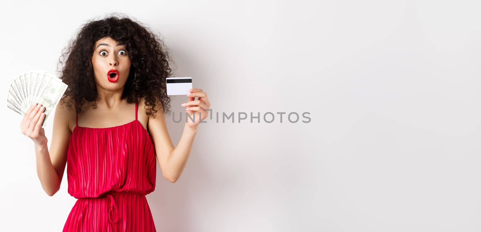 Impressed lady with curly hair and trendy red dress, saying wow, showing plastic credit card with cash, standing on white background.