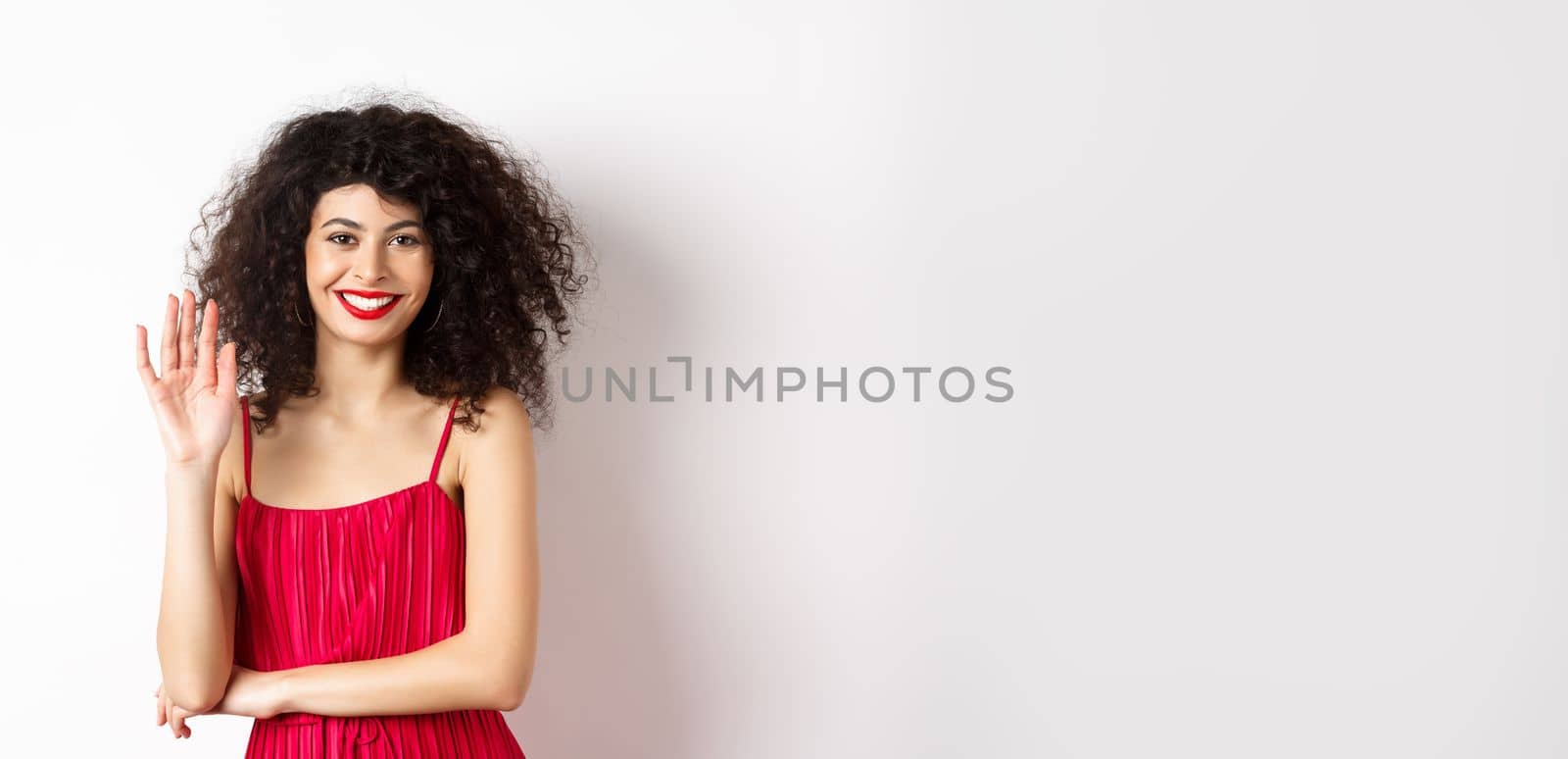 Cheerful elegant woman saying hello, waving hand and smiling at camera, greeting someone, standing in red dress on white background by Benzoix