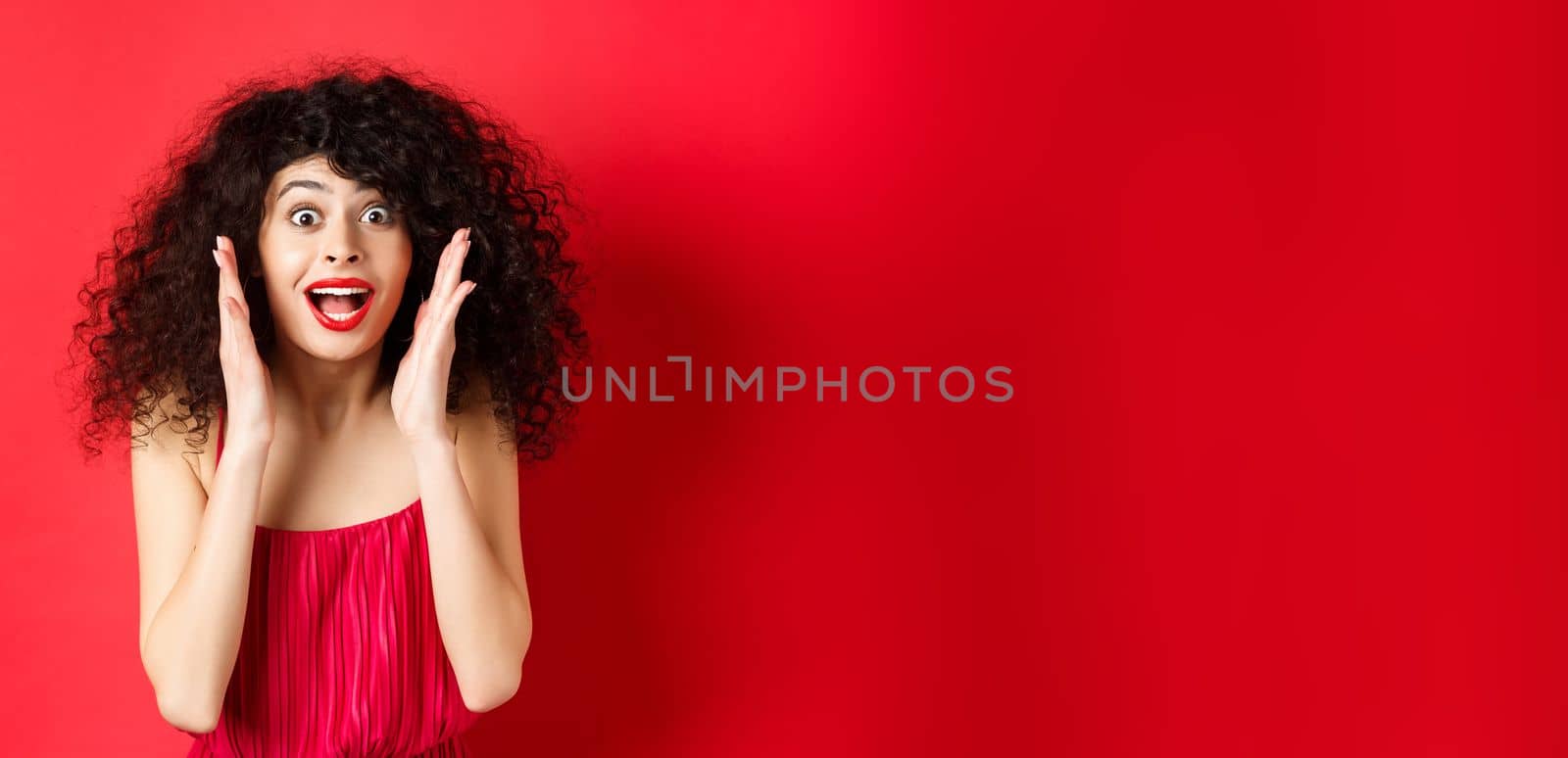 Close-up of surprised beautiful woman, screaming of amazement and looking at promo offer, standing in red dress and lipstick, studio background by Benzoix