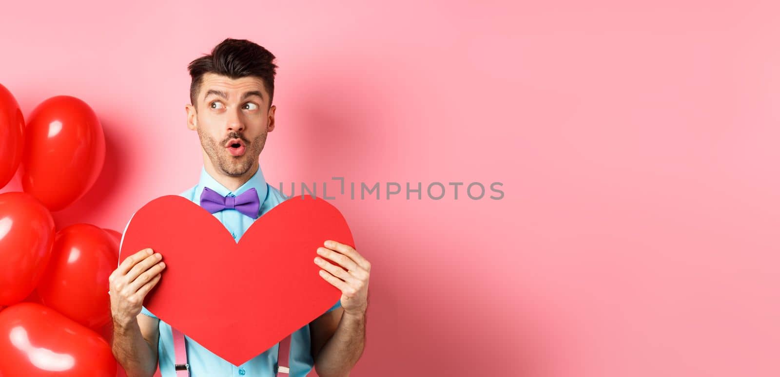 Valentines day concept. Cute young man looking left amused, showing red heart cutout and standing near balloons, pink background by Benzoix