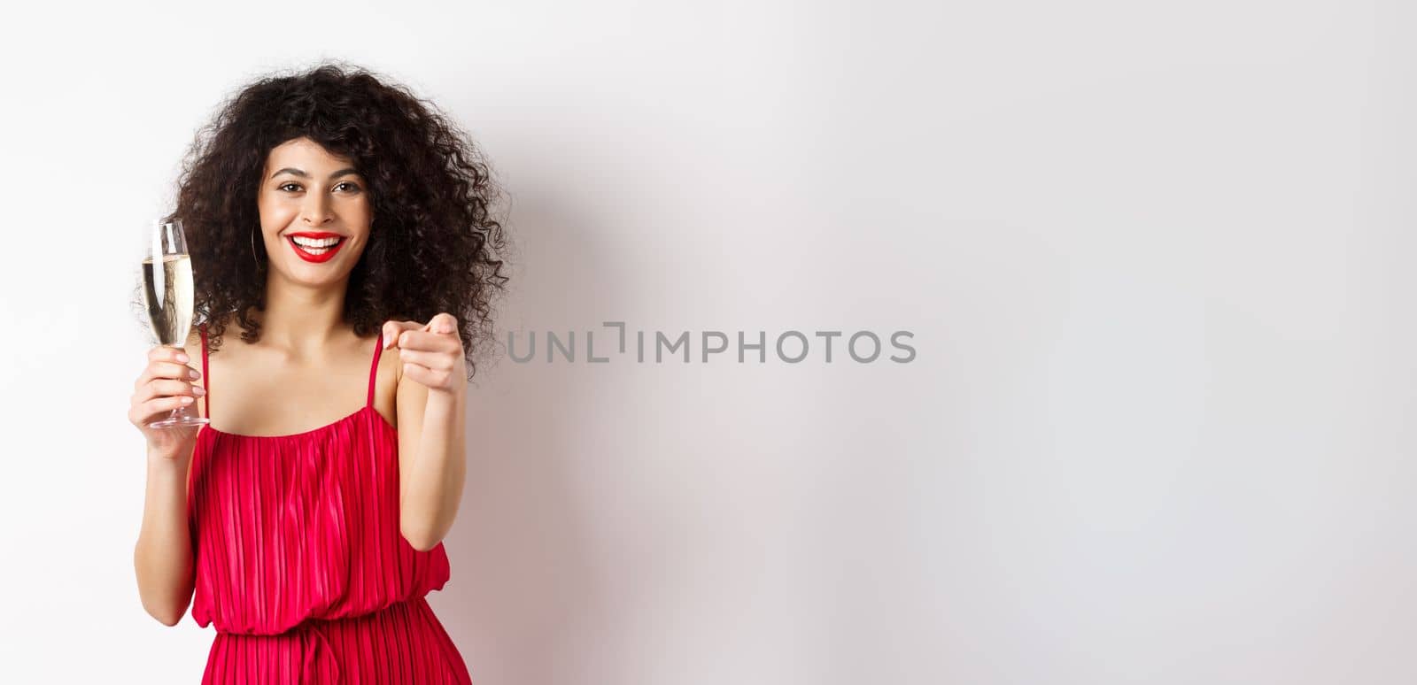 Beautiful curly woman in red dress, partying or having romantic date, holding glass of champagne and pointing at you, inviting person, standing on white background.