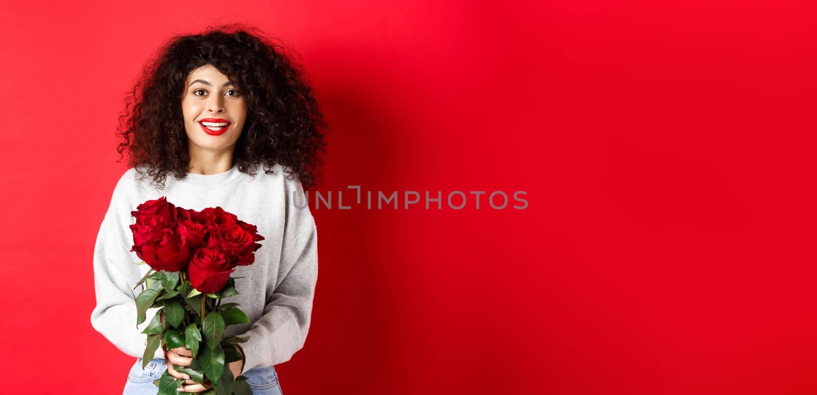 Romantic woman with curly hairstyle receive bouquet of red roses on Valentines day, looking tender at lover, standing on studio background by Benzoix