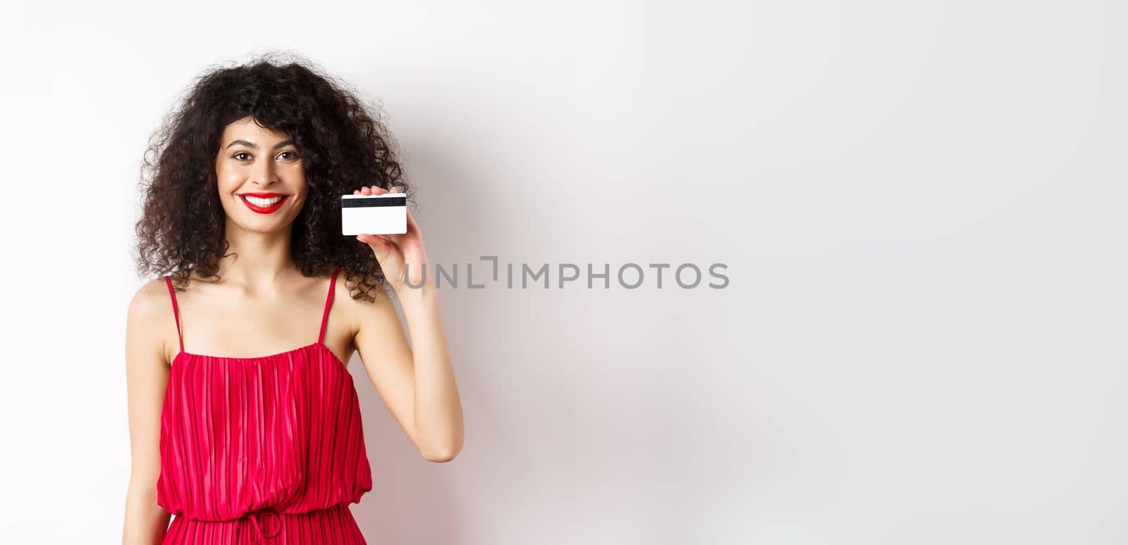 Beautiful woman in red trendy dress and makeup, showing plastic credit card and smiling, recommending offer, standing on white background.