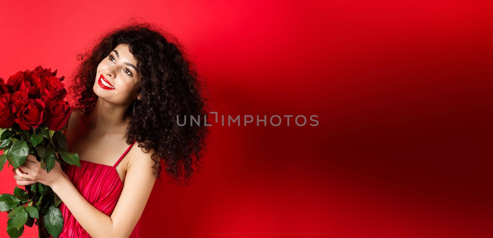 Happy beautiful woman in dress, holding flowers and smiling romantic, looking aside at logo, standing against red background.