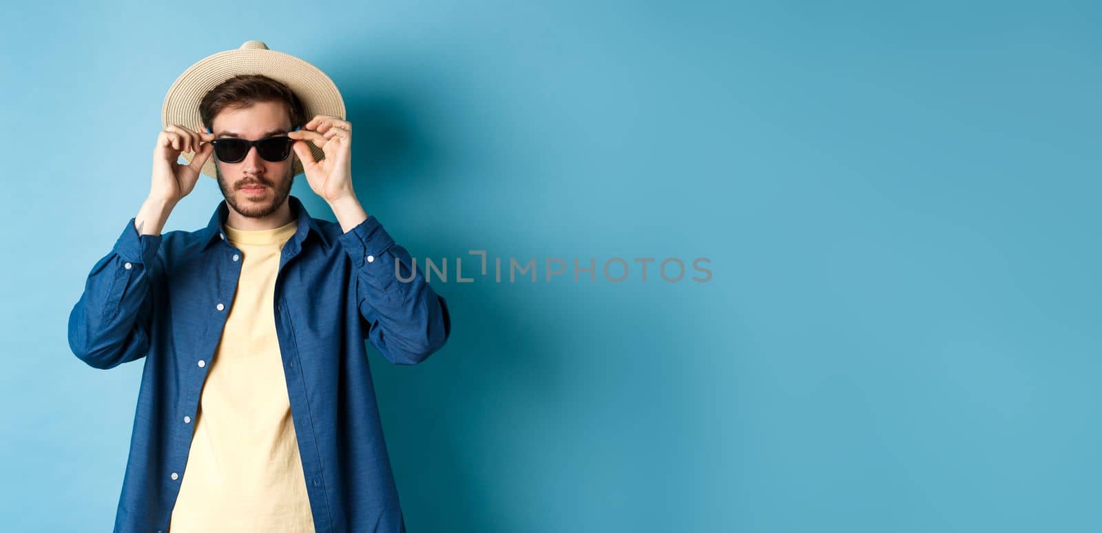 Handsome tourist in straw hat put on sunglasses on summer vacation, standing on blue background by Benzoix