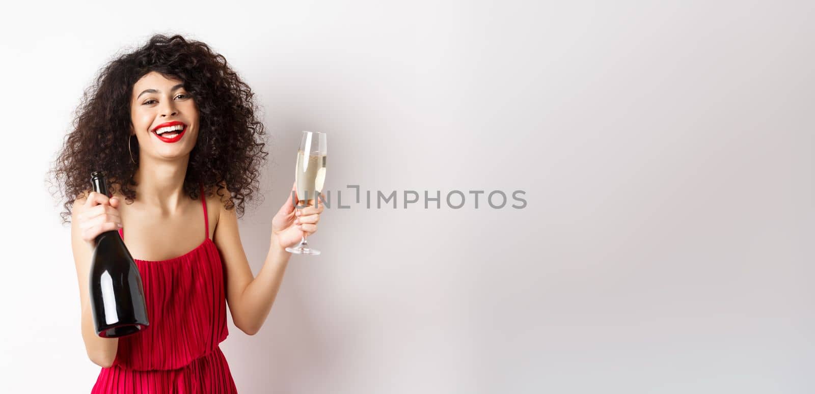 Happy party woman in red dress, laughing and holding bottle of champagne with glass, drinking and having fun, celebrating holiday, standing on white background by Benzoix