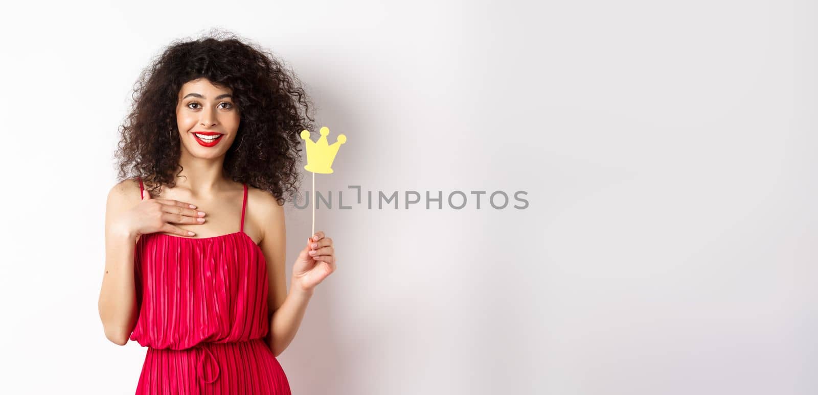 Confident pretty lady in red dress, holding queen crown on stick and looking excited, standing on white background.