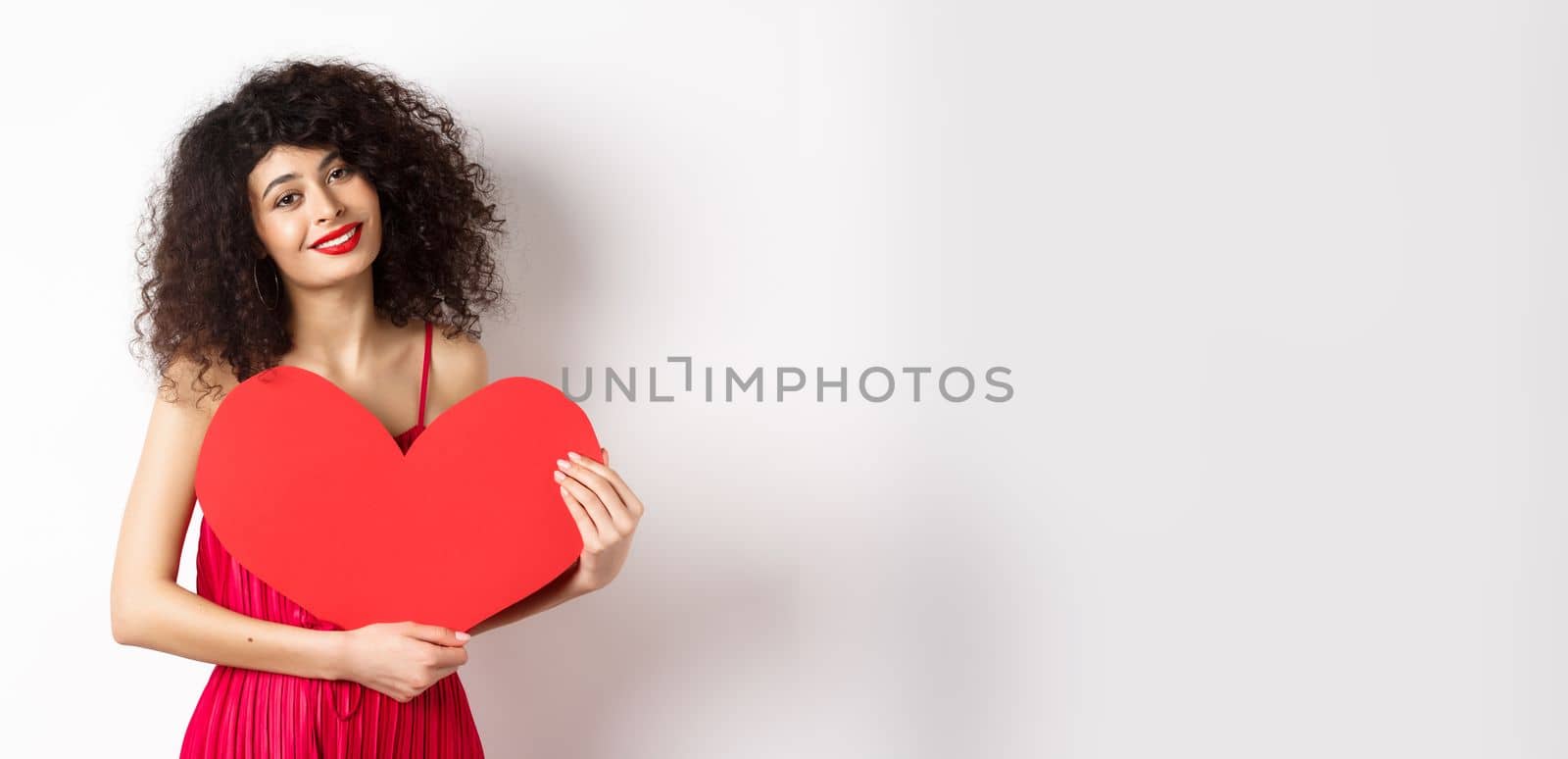 Romantic tender woman with curly hair, hugging big red heart and smiling, look with love, standing against white background.