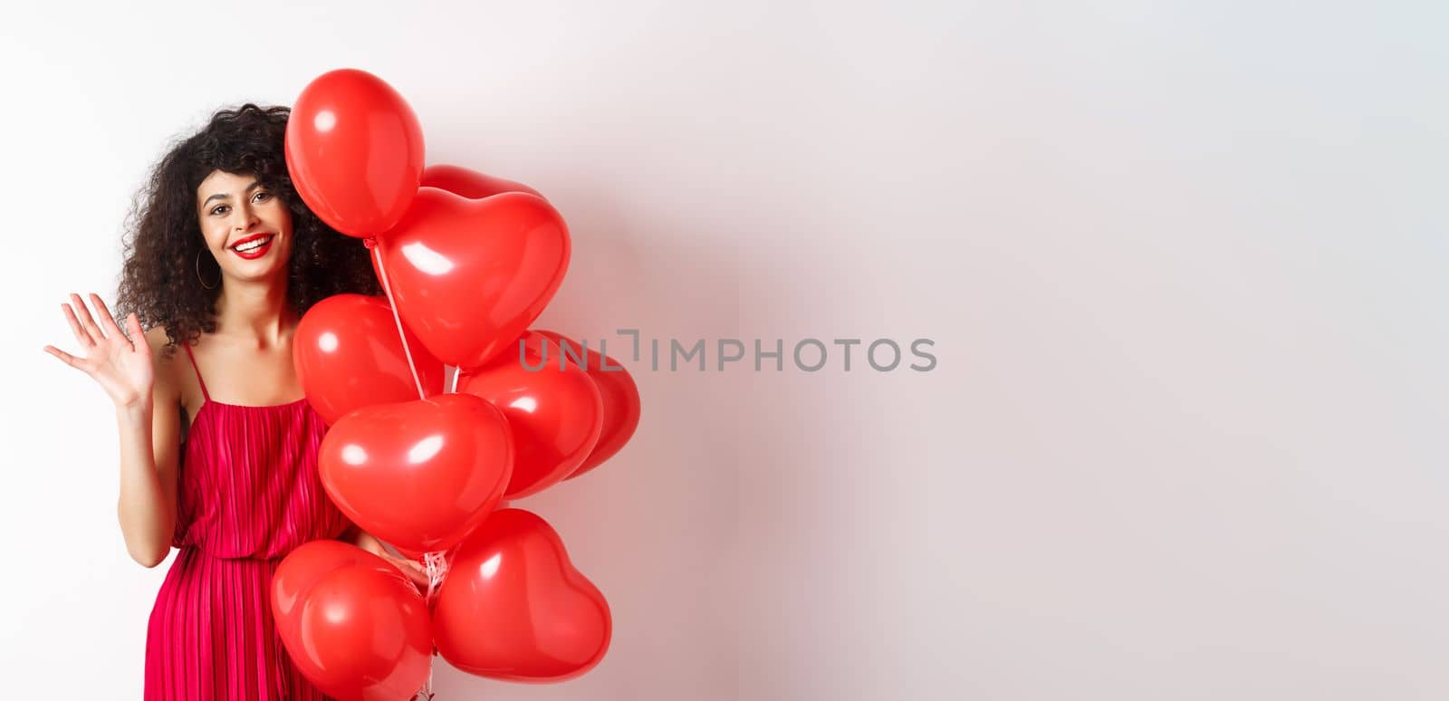 Beautiful lady with curly hair, standing near valentines holiday balloons and waving hand, saying hello, standing against white background.