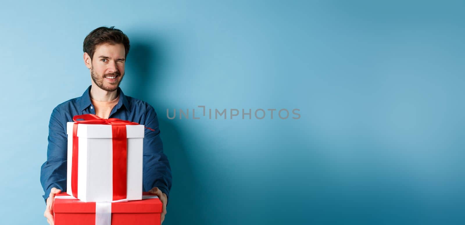 Handsome boyfriend winking at camera and wishing happy valentines day, extending hands with gift boxes, making romantic surprise, standing over blue background.